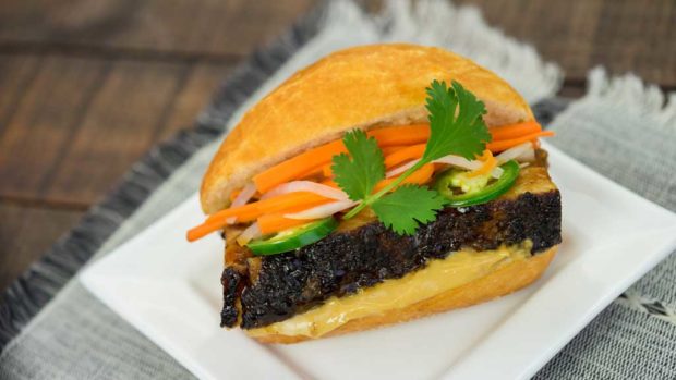 Black garlic soy-braised pork belly banh mi on a mini French roll with banh mi mayonnaise, garnished with pickled carrots and daikon, sliced jalapeño and cilantro sprig.