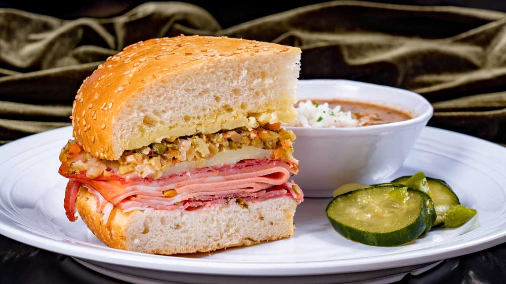 Muffuletta Sandwich – mortadella, salami, rosemary ham, cheddar, provolone and house-made olive relish on toasted New Orleans sesame seed bread, served with red beans & rice and house-made pickles.