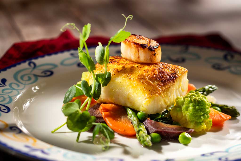 At Plaza de Coco, an array of artful dishes will transport guests to the picturesque town depicted in the Pixar Animation Studios film, “Coco.” Pan-Seared Adobo Spiced Sea Bass will be served alongside seared scallops, rainbow carrots, spinach, romanesco, peas and chimichurri sauce. (Kent Phillips, Photographer)